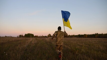 Soldier of ukrainian army running with raised blue-yellow banner on field at dusk. Young male military in uniform jogging with flag of Ukraine at meadow. Victory against russian aggression concept - 759084434