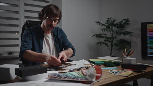 A stylish male architect is working on creating a project. The specialist is sitting at his workspace designing the interior. The worker is using blueprints, palettes, material samples, and textures.