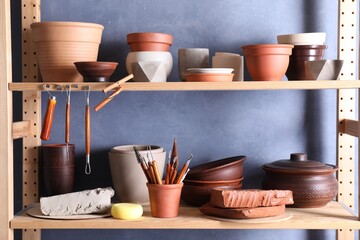 Set of different crafting tools and clay dishes on wooden rack in workshop