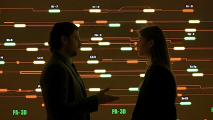 Technical support team in control room. Man and woman specialists discussing work, digital screen shows infrastructure monitoring infographic network data.