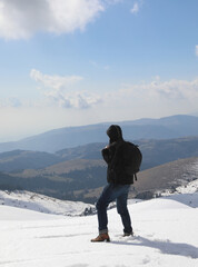 hiker with backpack and jacket looks at the view from the summit of the snow-capped mountain in the middle of winter - obrazy, fototapety, plakaty