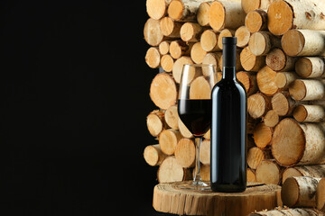 Stylish presentation of red wine in bottle and wineglass near wooden logs on black background,...