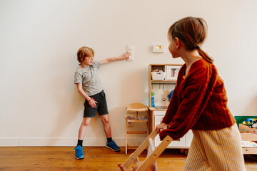 Child using the light switch indoors while playing with his sister