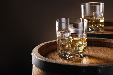 Whiskey with ice cubes in glasses on wooden barrels against dark background, closeup. Space for text