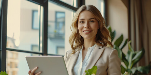 A Portrait of a happy businesswoman with a touchpad in the office looking at the camera standing beside a window and smiling. - obrazy, fototapety, plakaty