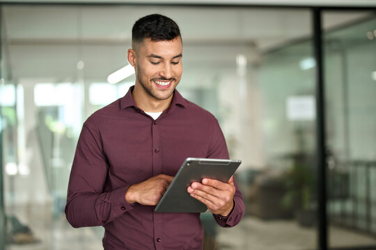 Happy hispanic businessman executive looking at tab device analyzing finance trading data working standing in office. Smiling busy young latin business man manager using tablet computer at work.