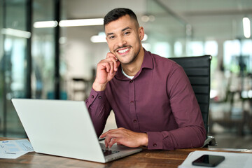 Happy young professional Latin business man looking at camera sitting at work desk using computer....