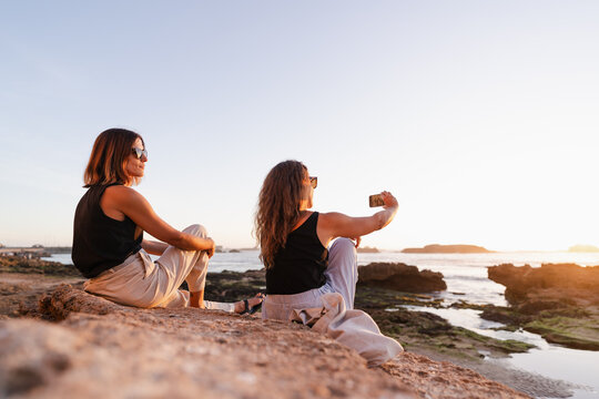 two women taking a selfie with their cell phone at sunset - Powered by Adobe