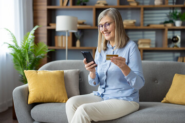 Cheerful mature woman sitting on the sofa, using her smartphone and holding a credit card, smiling...