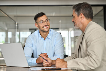 Young smiling professional latin business man working in corporate office with older executive manager using laptop technology having conversation on financial project strategy at work meeting.