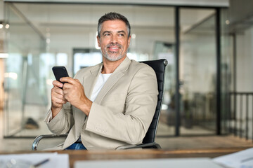 Happy busy middle aged business man investor wearing suit sitting in office using mobile cell phone. 50 years old businessman professional entrepreneur banker holding smartphone looking away at work.
