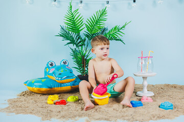 Summer photo zone for children. A little boy in shorts sits on the sand and plays with toys. Summer vacation.