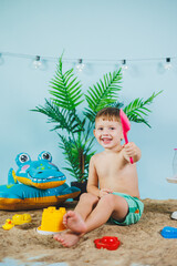 Summer photo zone for children. A little boy in shorts sits on the sand and plays with toys. Summer vacation.