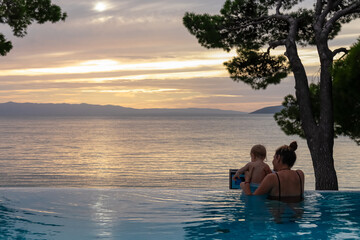 Loving mother with small toddler swimming in infinity pool during sunset at Makarska Riviera,...