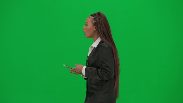 Female reporter isolated on chroma key green screen background. African American woman news host in suit walking and talking, holding tablet Side view.
