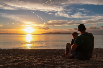 Father with small child enjoying romantic sunset at beach in town Makarska, Split-Dalmatia, Croatia, Europe. Coastline of Makarska Riviera, Adriatic Sea. Dreamlike atmosphere. Family vacation concept