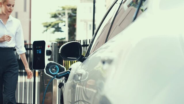 Businesswoman with shopping bag recharge her EV car display smart digital battery status hologram from charging station in car park or mall. Futuristic lifestyle and eco-friendly car. Peruse