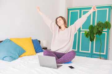 Happy woman sitting on the bed, working with computer and stretching hands at home - 759238844