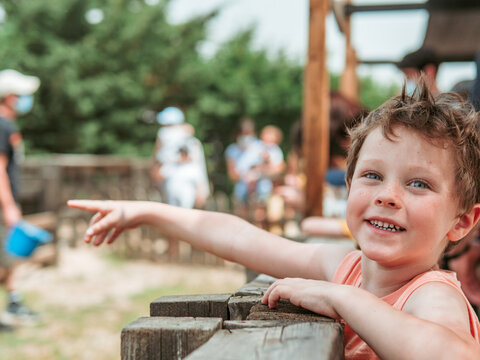 With sparkling eyes and a joyful smile, a young boy points excitedly at something off-camera at a bustling summer farm school