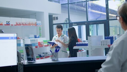 Obraz premium Asian woman in need of prescription medication informations in apothecary, asking cashier for help who redirects her to chemist checking pharmaceutical products inventory