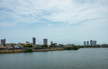 Fototapeta premium Cartagena, Colombia - July 25, 2023: Looking over bay water at old town from Puente Roman bridge. Historic reinforced shore ramparts under light blue cloudscape. Few tall buildings