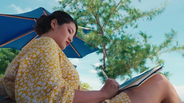 Korean female artist sitting outdoors and drawing summer landscape. Asian woman wearing yellow dress painting watercolor sketch on plain-air. Happy summertime. Vacation leisure. Holidays in tropics. 