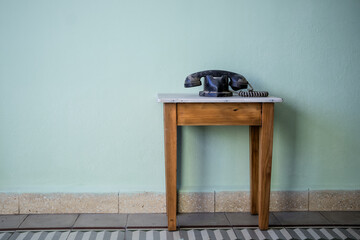 vintage black telephone covered in dust reststing on a small wooden table against a plain wall. - obrazy, fototapety, plakaty