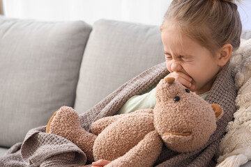Sick little girl with toy bear sneezing on sofa at home, closeup