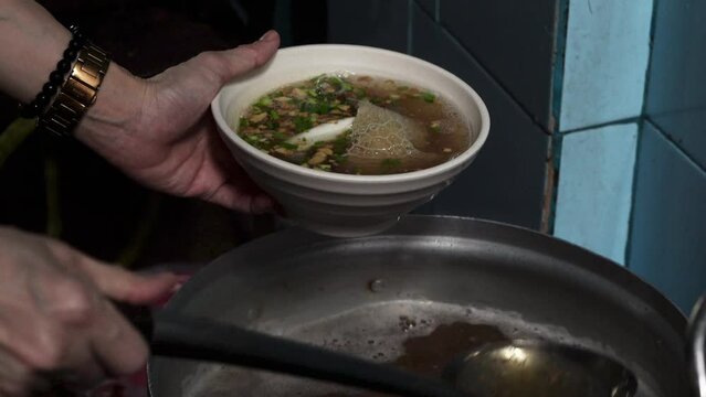 A close-up of hands of a person pouring meat and offal soup from a metal pot into a white ceramic bowl using a ladle.