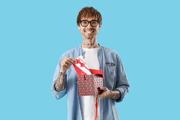 Young tattooed man with gift voucher in box on blue background