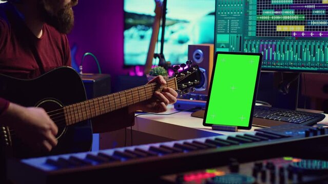 Male artist learning to play guitar on online lesson tutorial, practicing acoustic instrument and looking at phone with greenscreen display. Music producer doing rehearsal guitar play. Camera A.