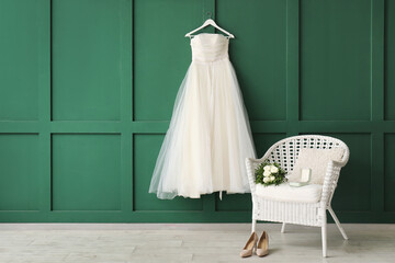 Beautiful wedding dress on green wall, wedding shoes on floor and bridal bouquet on white chair in room