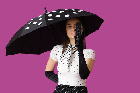 Young woman with umbrella and retro movie camera on purple background