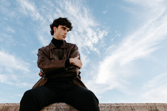 Stylish young man sitting on stone border against blue sky