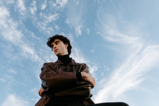Young confident man in leather jacket against blue sky with clouds