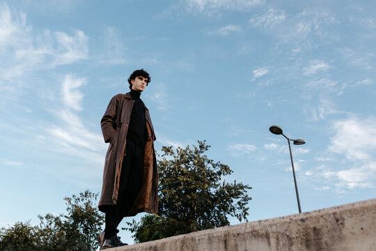 Stylish young man walking on stone border against blue sky