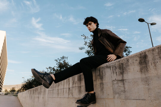Trendy young man sitting on stone border against blue sky in street