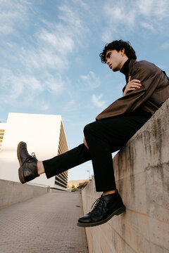 Stylish man sitting on stone border in city street against blue sky