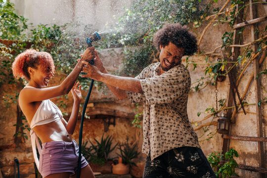 Black couple teasing each other playing with a water hose in summer