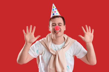 Young man in party hat and clown nose showing tongue on red background. April Fool's Day celebration