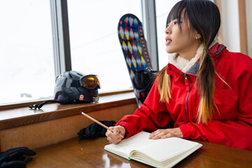 Asian woman writing and drawing in diary book during practicing snowboard on snowy mountain at ski...