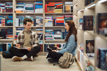 Young man and woman sitting on floor discussing with books in library.