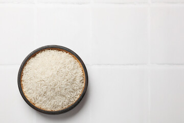 Raw basmati rice in bowl on white tiled table, top view. Space for text