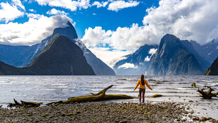 Happy girl in yellow jacket admires famous Milford Sound fiord and mighty mountains densely covered with vegetation in Fiordland National Park, Southland, New Zealand
