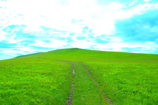Mystical scenery from Piani di Ragnolo with a beaten path traversing the vast green expanses and heading to the left, a pole on the way and a vision of peaks in the background under a cloudy azure sky