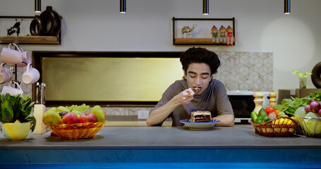 Asian Indian Hindu happy teen boy sitting modern kitchen hold spoon eating sweet tasty pastry cake tasting food snacks chewing bite indoor house cute gen z male enjoy fun joy holiday home