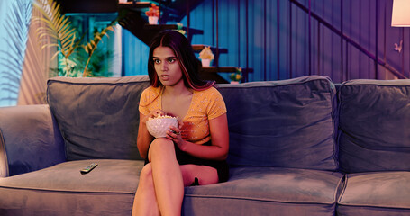 Obraz premium Indian Asian Hindu gen z young woman sitting sofa couch watch horror scary movie indoor home scared adult afraid girl hold bowl eat snacks look series film on TV enjoy fun joy late night room house