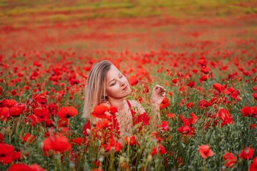 Naklejka premium Happy woman in a red dress in a beautiful large poppy field. Blond sits in a red dress, posing on a large field of red poppies