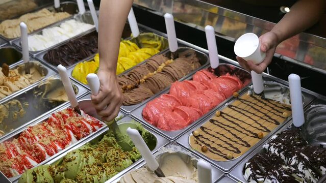 Ice cream seller carefully spoons portion of flavored ice cream from tray into paper cup with spatula. Multi-colored ice cream scoops look delicious. Sweet ice cream treat is welcome treat on hot day.