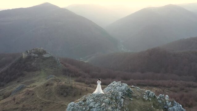 Slow motion aerial view of bride in white wedding dress standing on a cliff